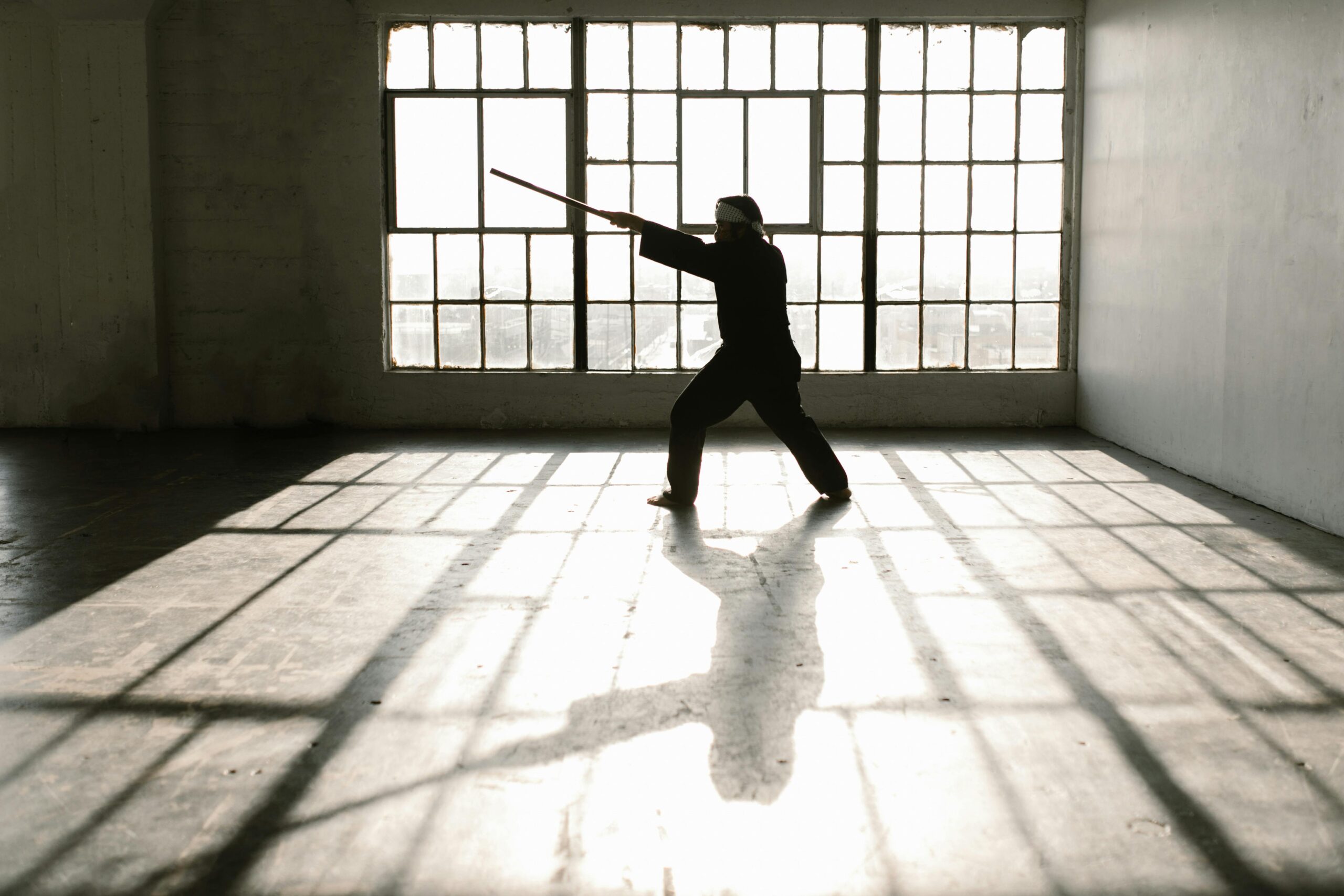 Silhouette of martial artist practicing with a staff in a sunlit studio.